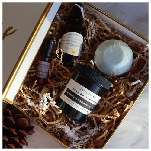 A gift set containing a jar of shea butter, a container of scrub, a shower steamer, and a bottle of oil, all displayed in a box with shredded paper bedding.